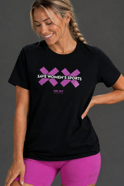 A woman wearing a black short-sleeve t-shirt with a purple graphic that reads 'SAVE WOMEN'S SPORTS' and 'XX-XY SPORTS' underneath, paired with bright pink athletic shorts, against a dark gray studio background. || Bright Fuchsia