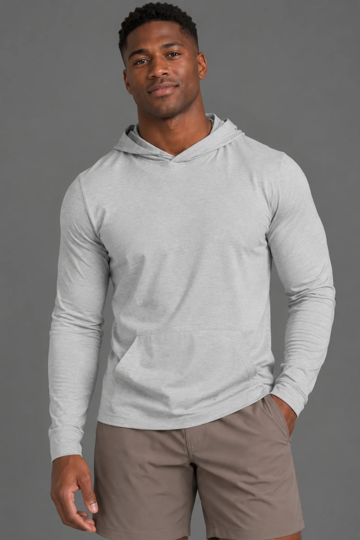 Black male model wearing a light gray performance hoodie and brown athletic shorts with one hand in his pocket. || Light Heather Gray - 1