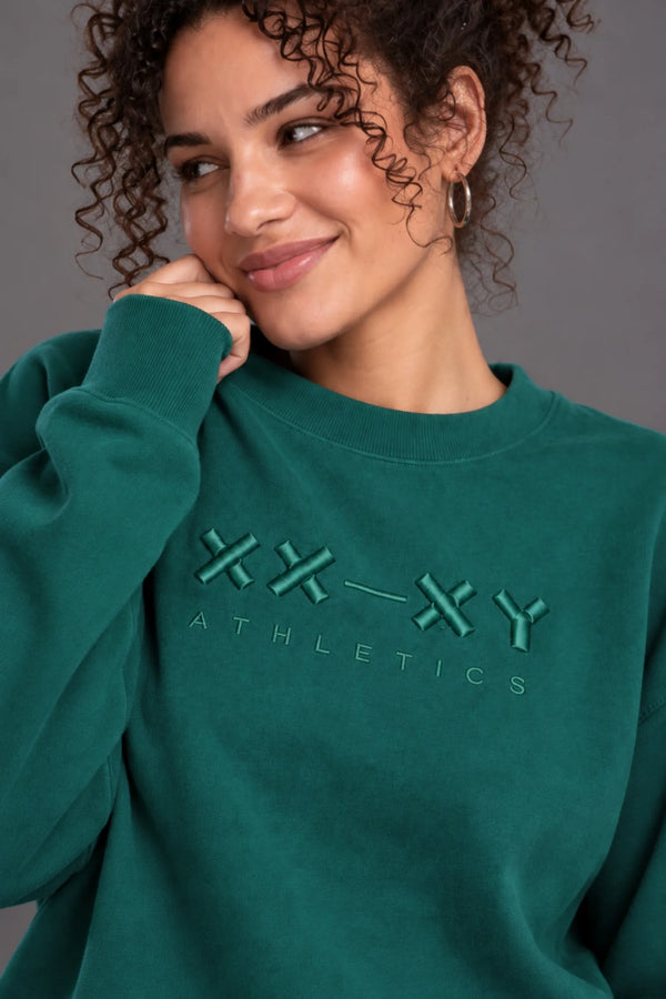 Close-up of a woman wearing a teal XX-XY Athletics crewneck sweatshirt, highlighting the tonal embroidered chest logo. || Pine