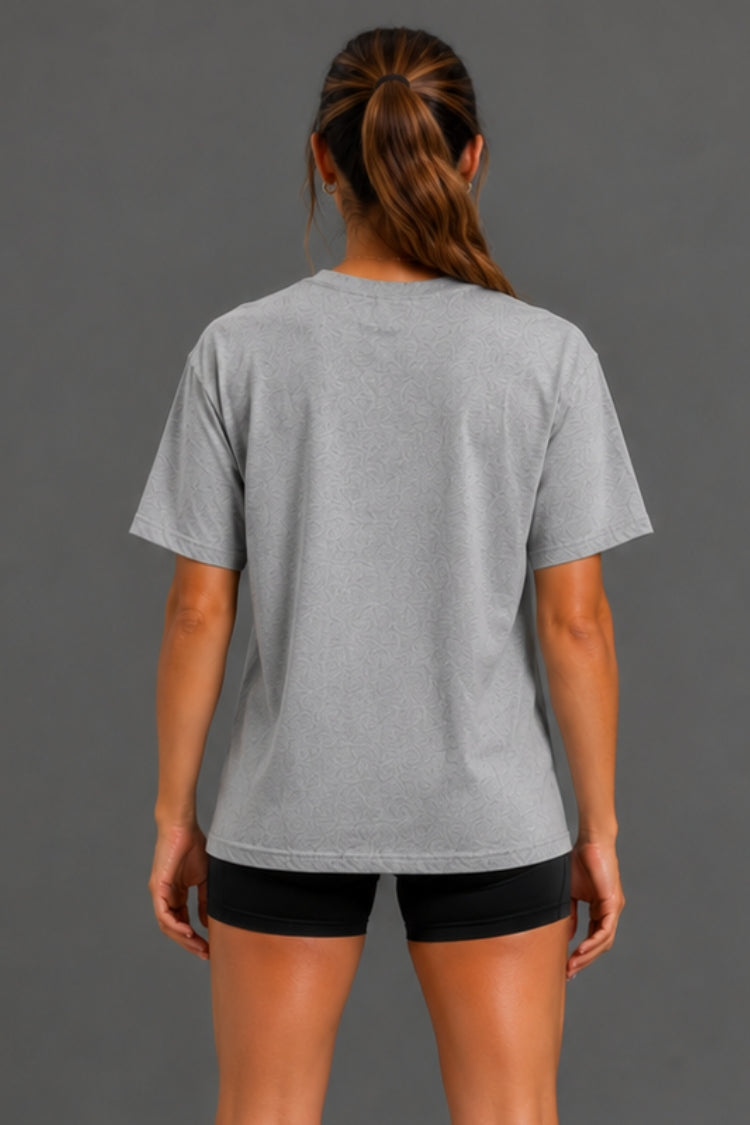 A woman viewed from the back wearing a light gray short-sleeve crewneck t-shirt and black athletic shorts, set against a solid dark gray studio background.
