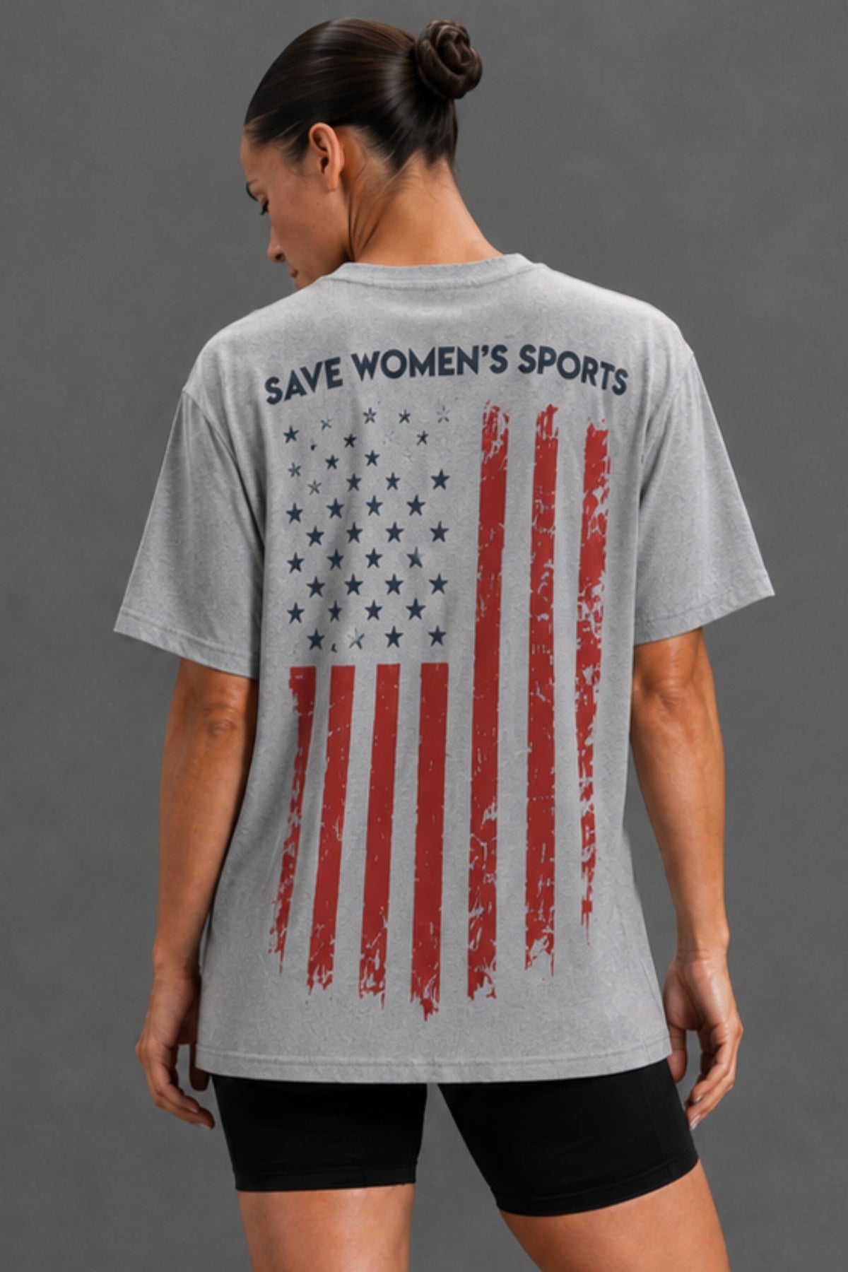 A woman shown from the back wearing a heather gray graphic T-shirt with a distressed American flag print and the text 'SAVE WOMEN'S SPORTS' across the upper back, paired with black shorts against a plain gray studio background. || Heather Gray