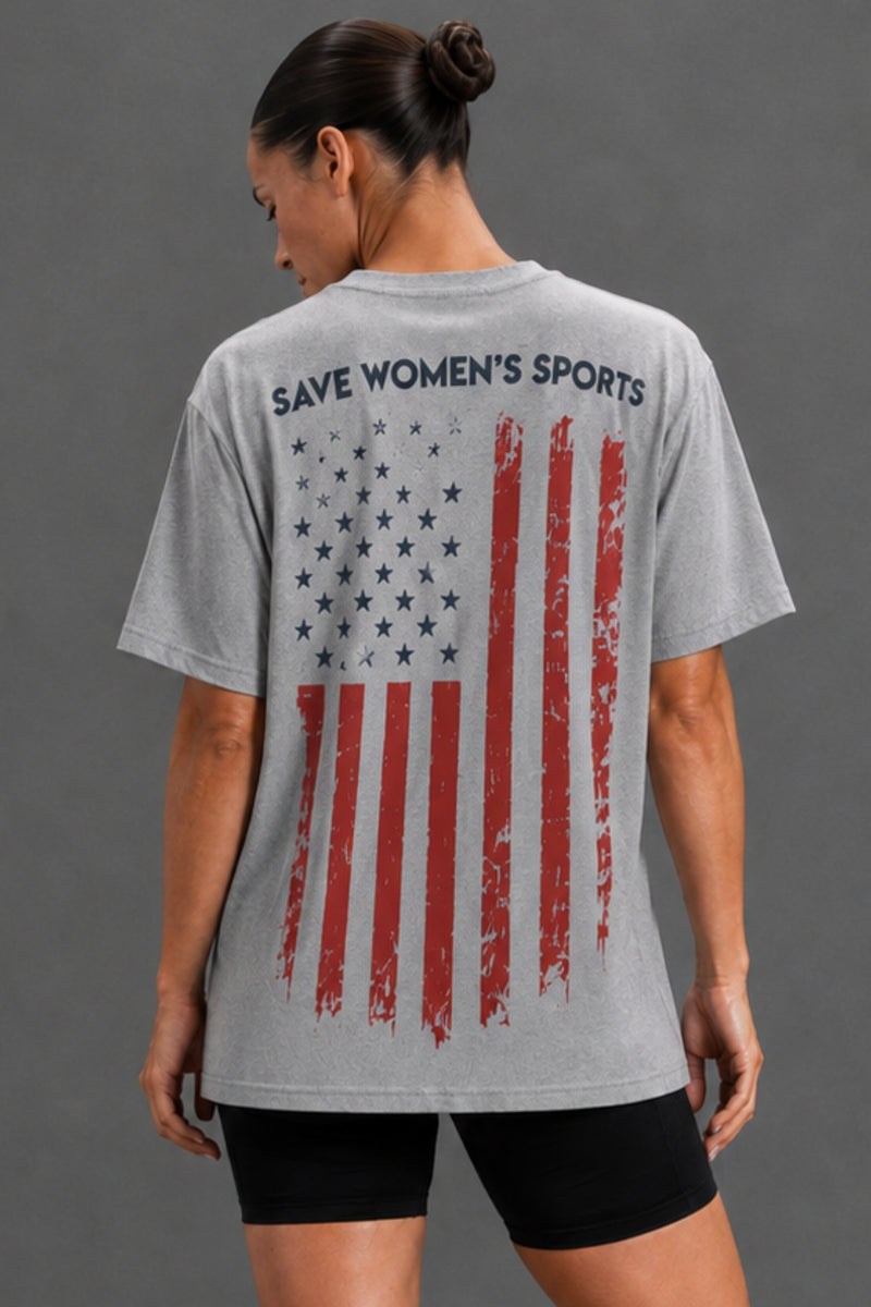 A woman shown from the back wearing a heather gray graphic T-shirt with a distressed American flag print and the text 'SAVE WOMEN'S SPORTS' across the upper back, paired with black shorts against a plain gray studio background. || Heather Gray