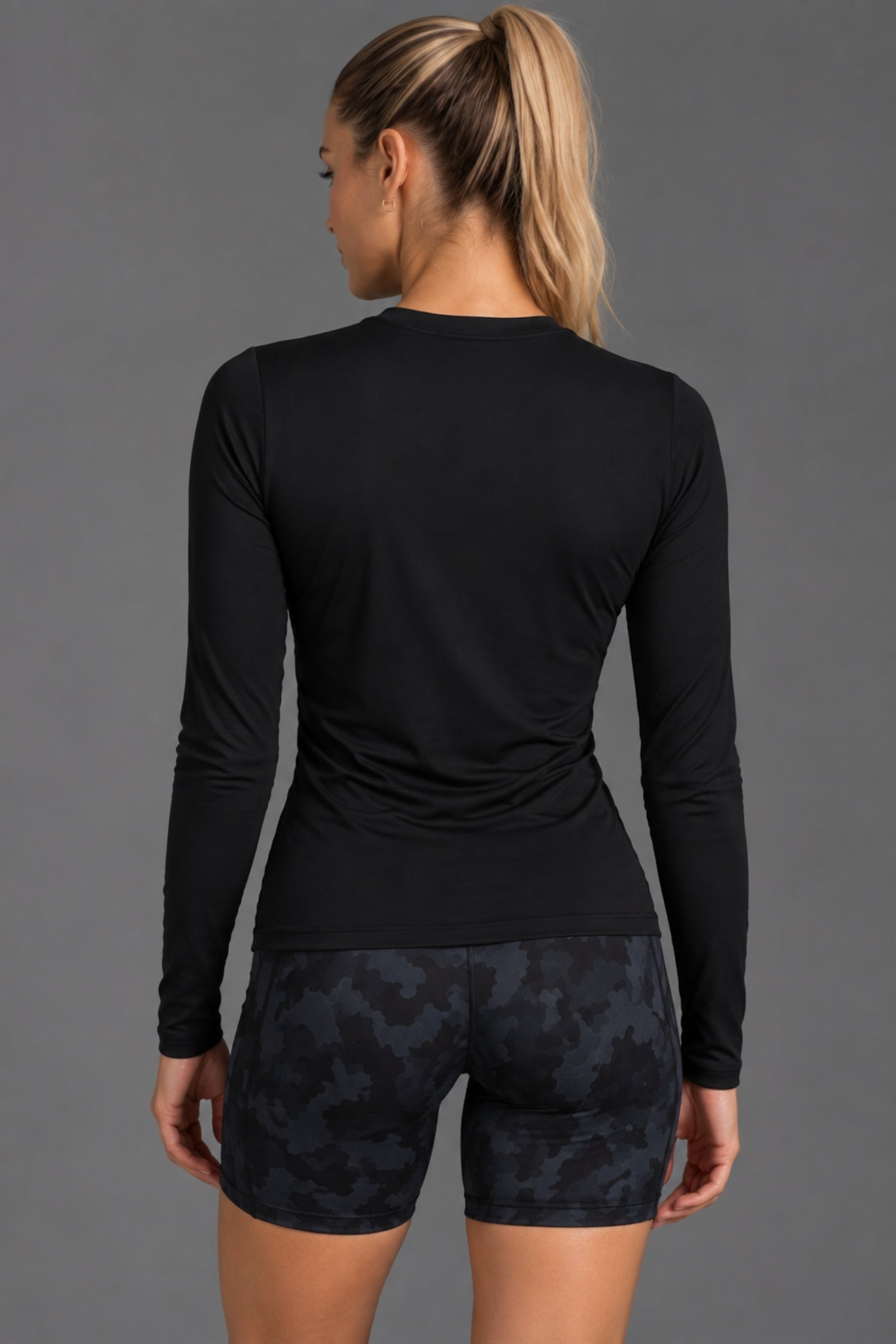 A woman shown from the back wearing a fitted black long-sleeve athletic top and dark patterned shorts against a plain gray studio background. || Black