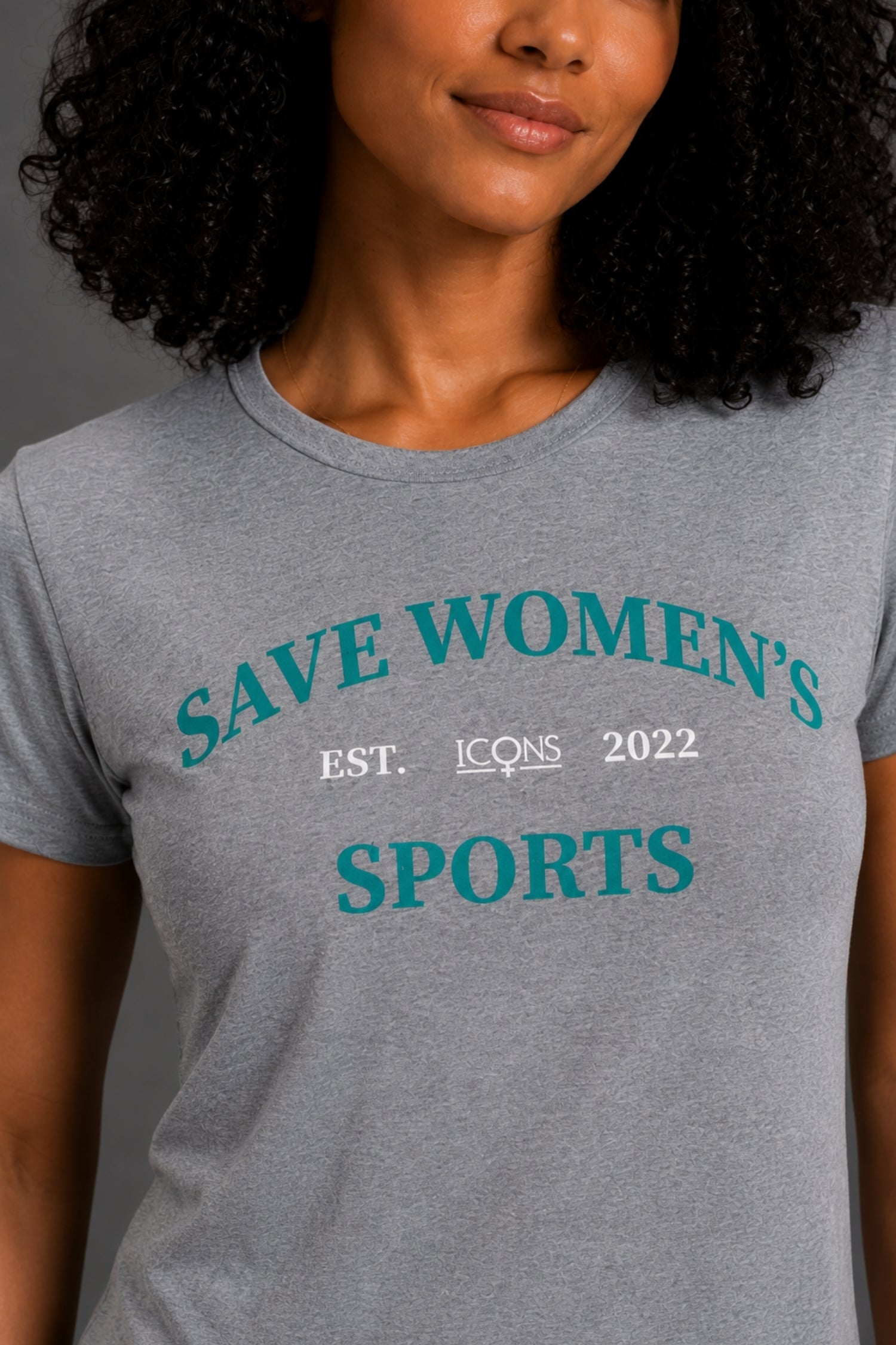 A smiling woman with curly black hair wearing a heather gray graphic t-shirt with teal text reading 'SAVE WOMEN'S SPORTS' and black pants, standing against a solid gray studio background. || Heather Gray