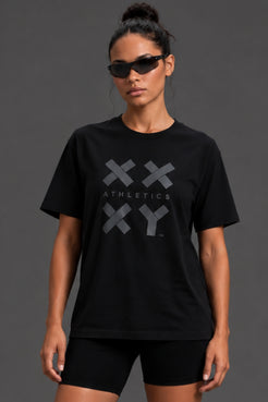 A woman modeling a black short-sleeve athletic T-shirt with a large gray XXY Athletics logo on the front, paired with black bike shorts against a dark gray studio background. || Black Tonal