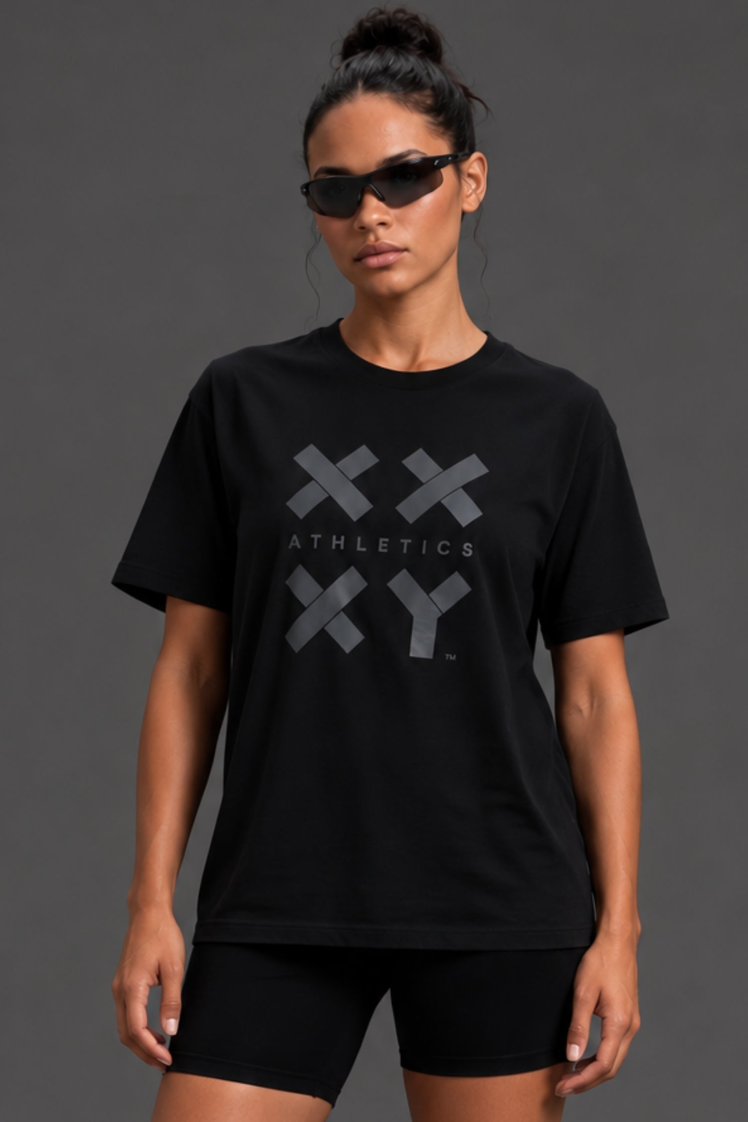 A woman modeling a black short-sleeve athletic T-shirt with a large gray XXY Athletics logo on the front, paired with black bike shorts against a dark gray studio background. || Black Tonal