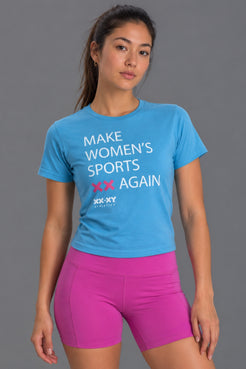 Female wearing a blue  "Make Women's Sports XX Again" t-shirt styled with pink bike shorts shot on a studio gray background. || Light Blue