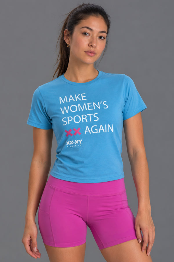 Female wearing a blue  "Make Women's Sports XX Again" t-shirt styled with pink bike shorts shot on a studio gray background. || Light Blue