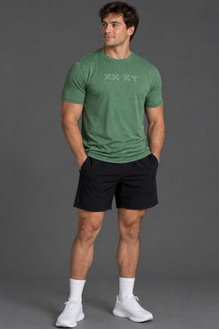 A man modeling a green short-sleeve athletic t-shirt with a subtle chest logo, paired with black shorts, white socks, and white sneakers, set against a solid gray studio background. || Hunter Green Heather