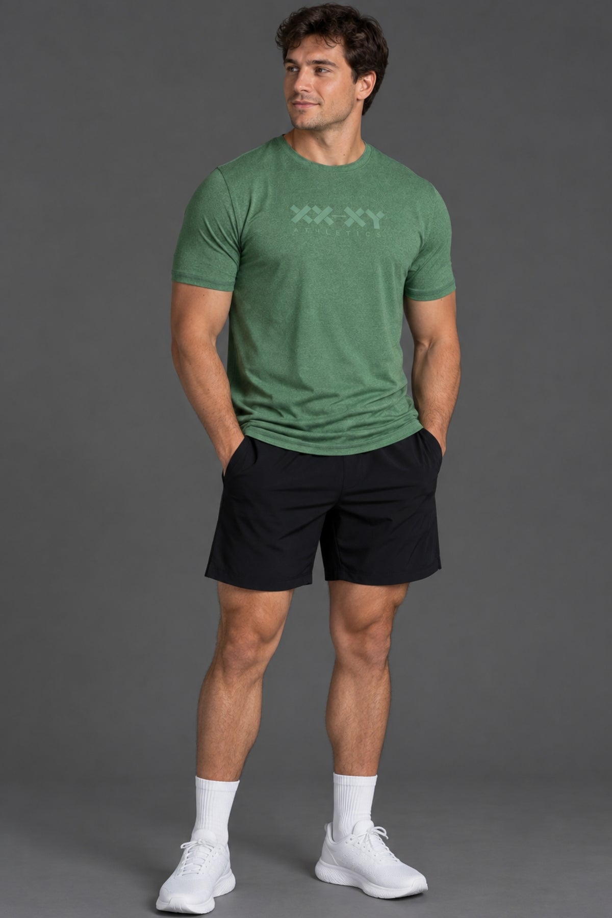A man modeling a green short-sleeve athletic t-shirt with a subtle chest logo, paired with black shorts, white socks, and white sneakers, set against a solid gray studio background. || Hunter Green Heather