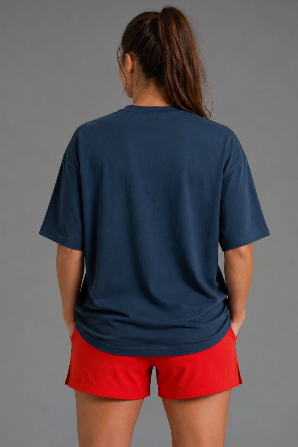 A woman shown from the back wearing an oversized navy blue t-shirt and bright red athletic shorts against a solid gray studio background.