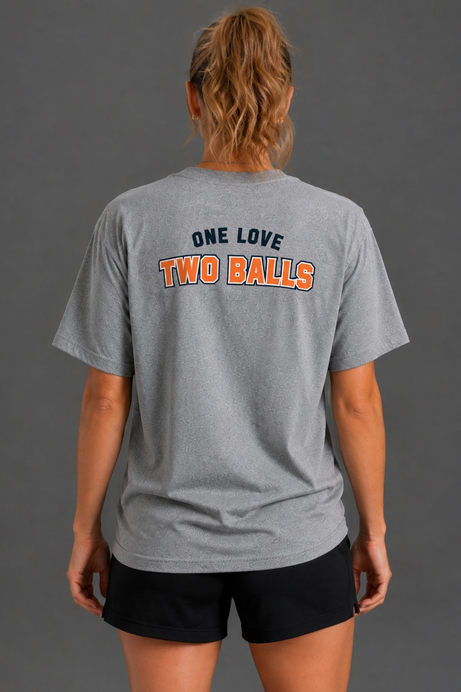 A woman viewed from behind wearing a heather gray t-shirt with the text 'ONE LOVE TWO BALLS' printed on the back in navy and orange, paired with black athletic shorts against a dark gray studio background. || Heather Gray