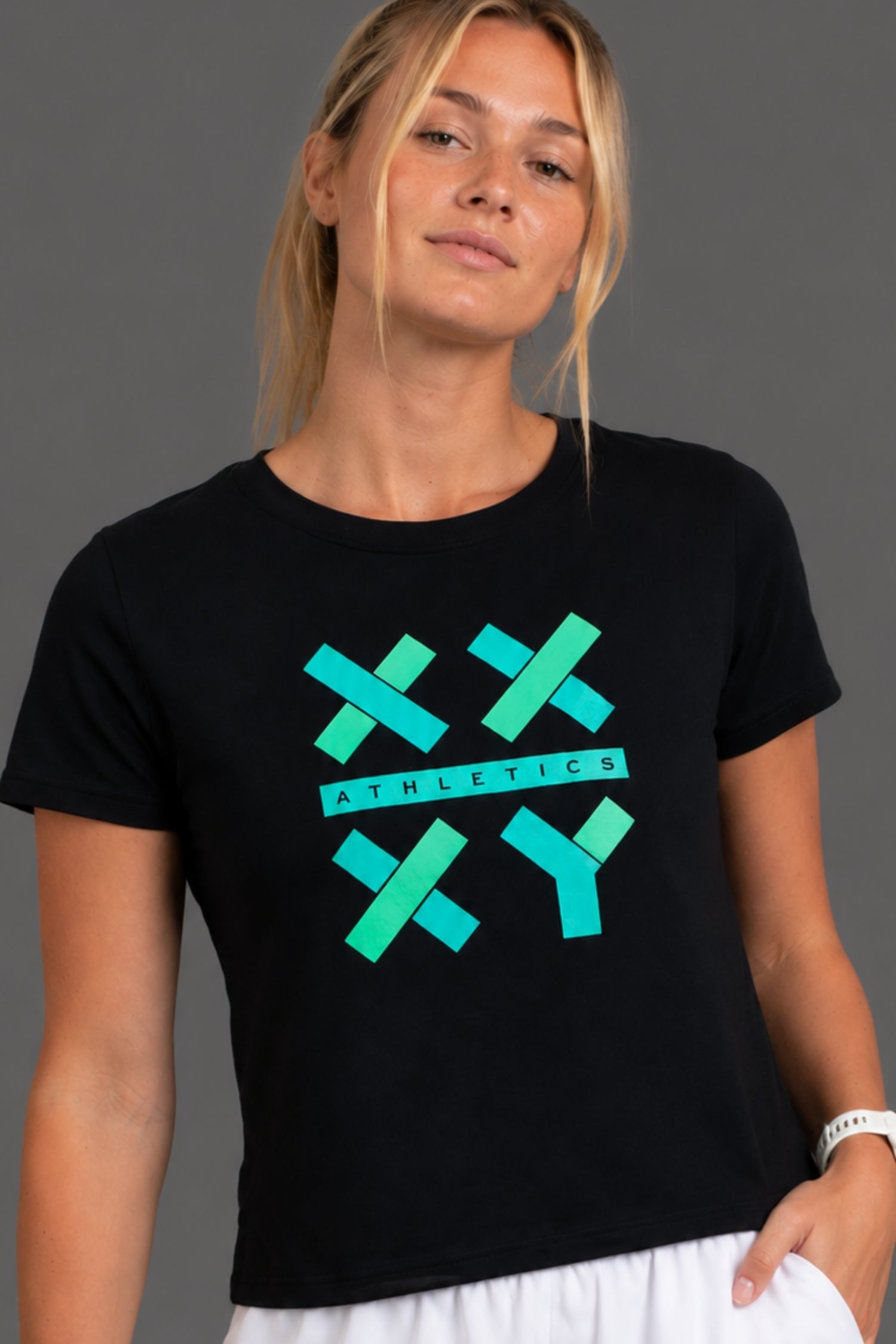 A woman wearing a black short-sleeve crewneck t-shirt with a teal and green athletic graphic on the front, paired with white pants, against a solid gray studio background. || Black