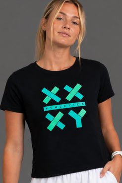 A woman wearing a black short-sleeve crewneck t-shirt with a teal and green athletic graphic on the front, paired with white pants, against a solid gray studio background. || Black