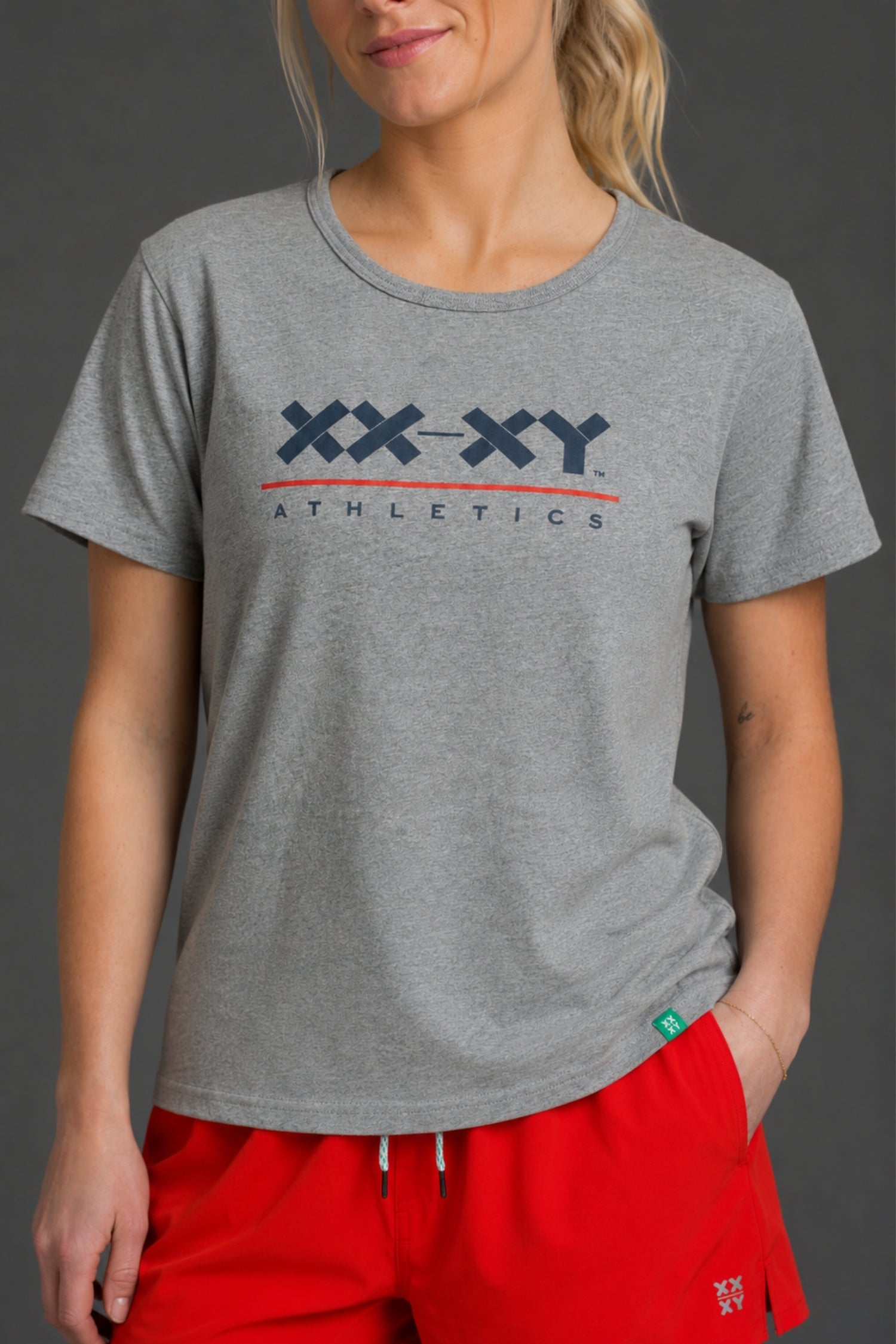 A woman wearing a heather gray crewneck t-shirt with a navy and red 'XX-XY ATHLETICS' logo on the chest, paired with bright red athletic shorts, against a dark gray studio background. || Light Heather Gray