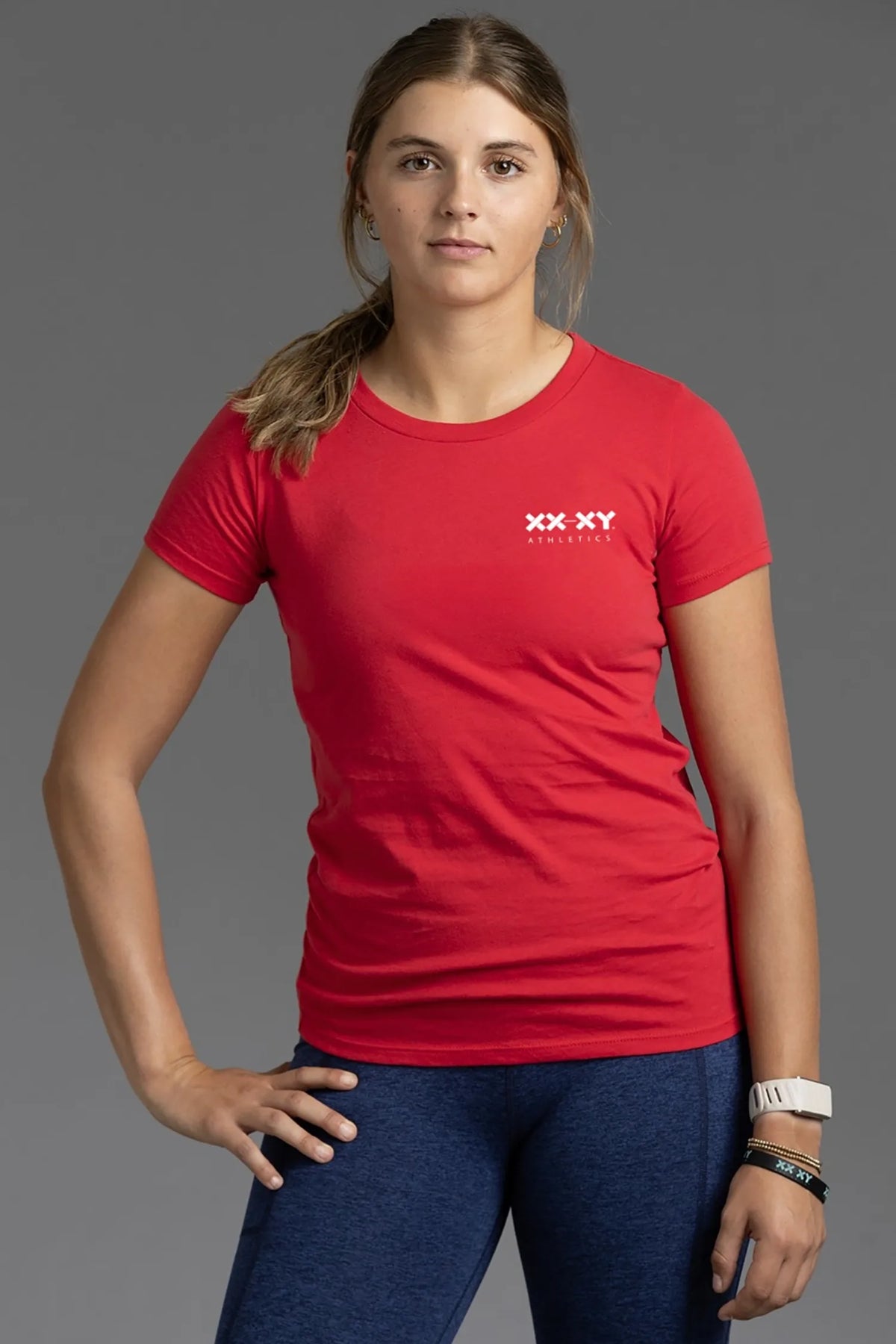 Female model wearing a red XX-XY Athletics crew neck T-shirt with a white chest logo and navy leggings, standing against a gray studio background. || Red