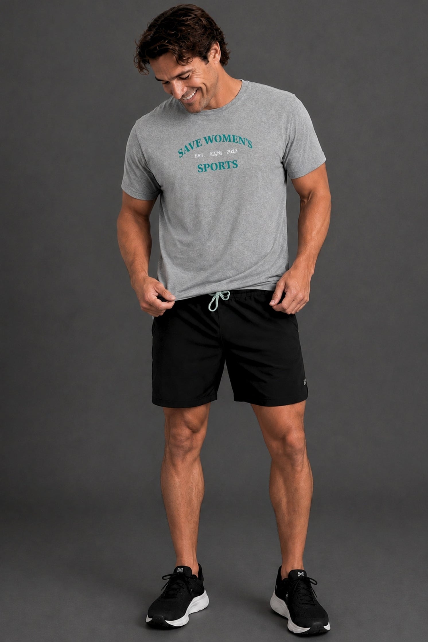 A man wearing a heather gray t-shirt with 'SAVE WOMEN'S SPORTS' printed in teal, paired with black athletic shorts and black sneakers, standing against a dark gray studio background. || Heather Gray