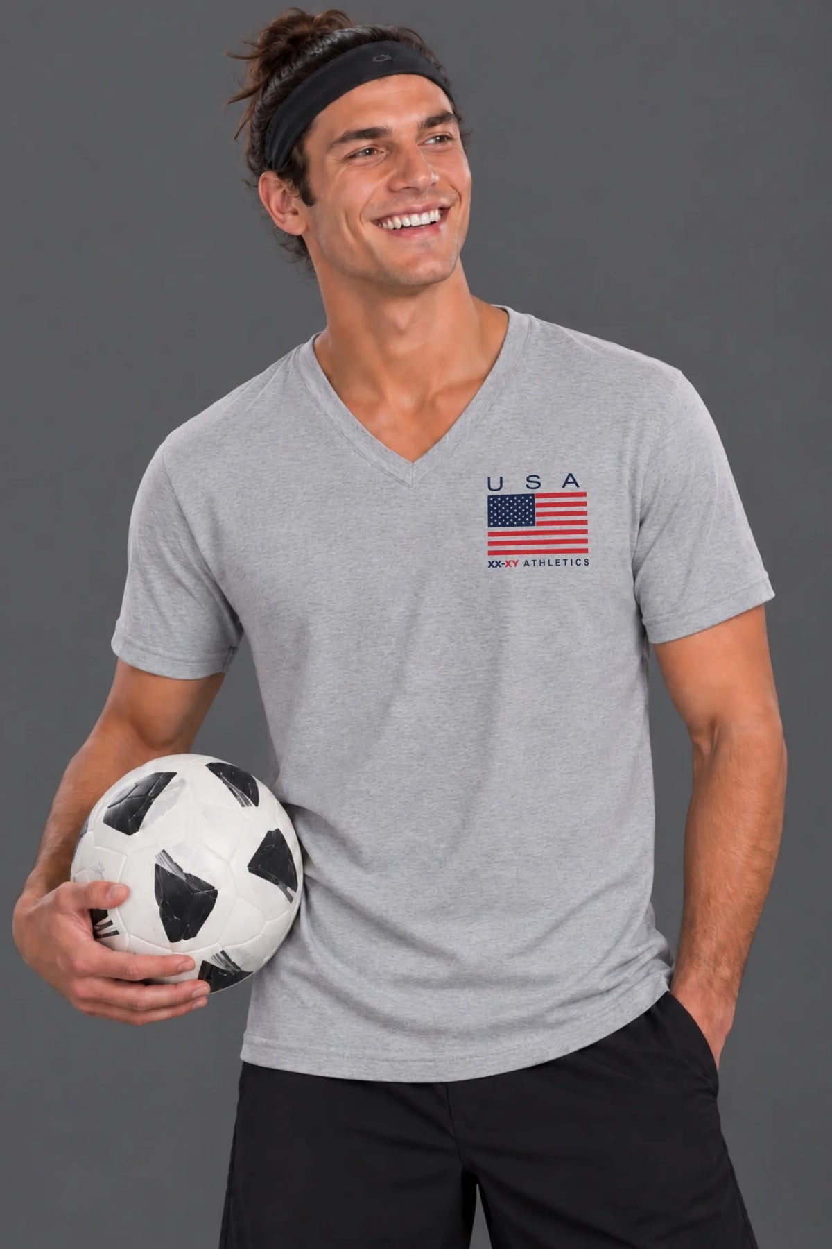 Male model standing against a gray studio background wearing the heather gray XX-XY Athletics USA Flag V-Neck Tee, black athletic shorts, white socks, and soccer cleats.