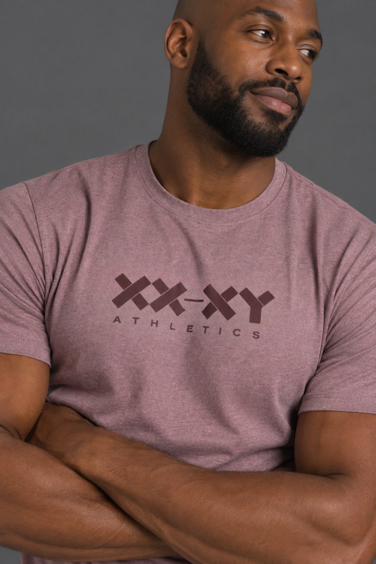 A close-up of a muscular man wearing a heathered mauve-pink t-shirt with a dark brown 'XX-XY ATHLETICS' logo printed on the chest, set against a solid gray background.