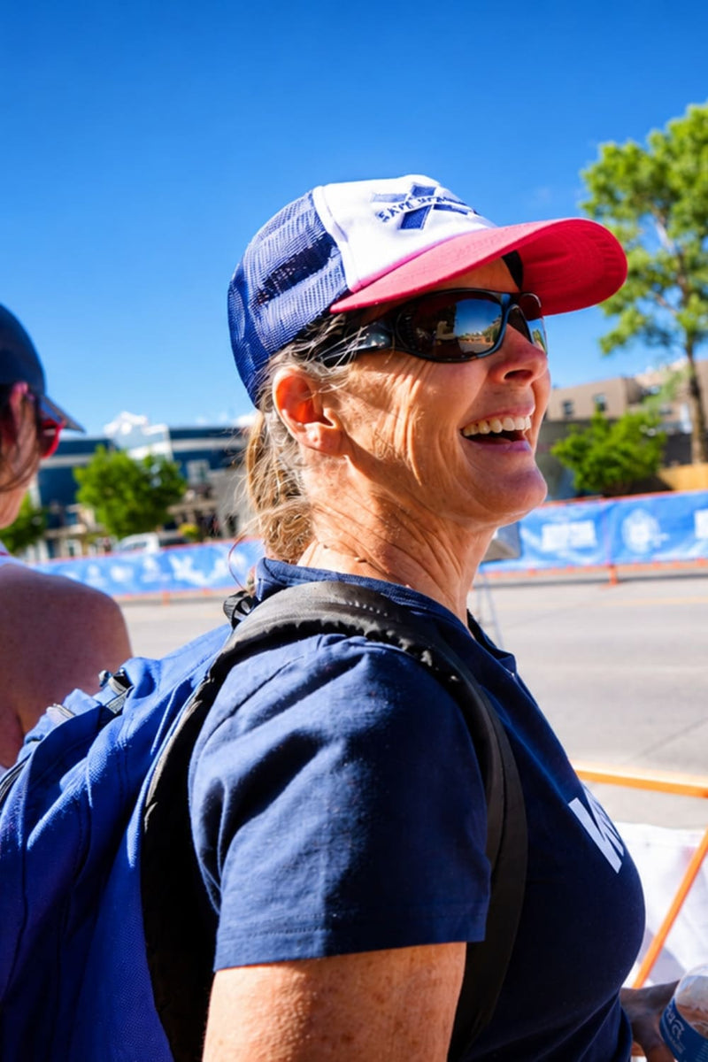 A woman outdoors wearing a red, white, and blue trucker hat, dark sunglasses, a navy blue t-shirt, and a blue backpack, smiling in a sunny outdoor setting.