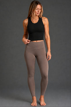 A woman modeling high-waisted, ankle-length leggings in a taupe brown color, paired with a black sports bra, against a solid dark gray studio background. || Black