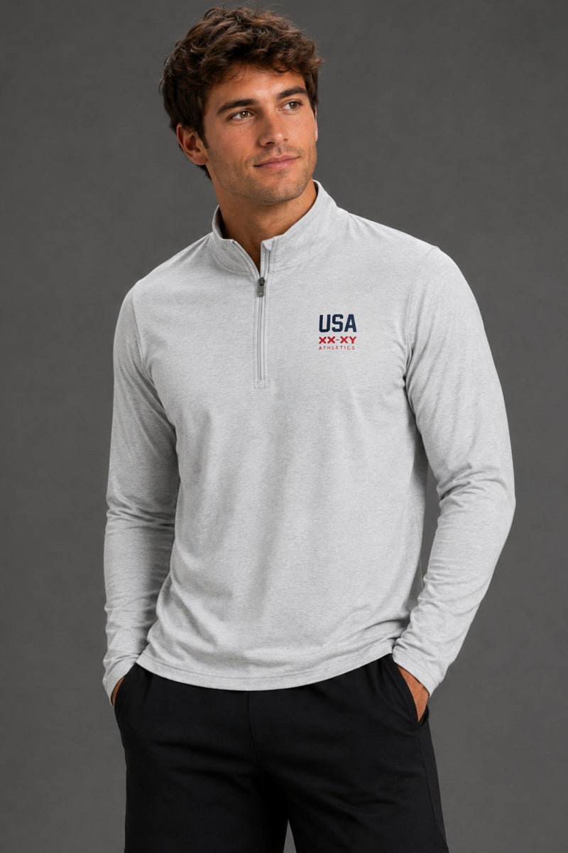 A man wearing a light gray heather long-sleeve quarter-zip pullover with a 'USA HOCKEY' logo on the left chest, paired with black pants, against a dark gray studio background. || Light Heather Gray - 1