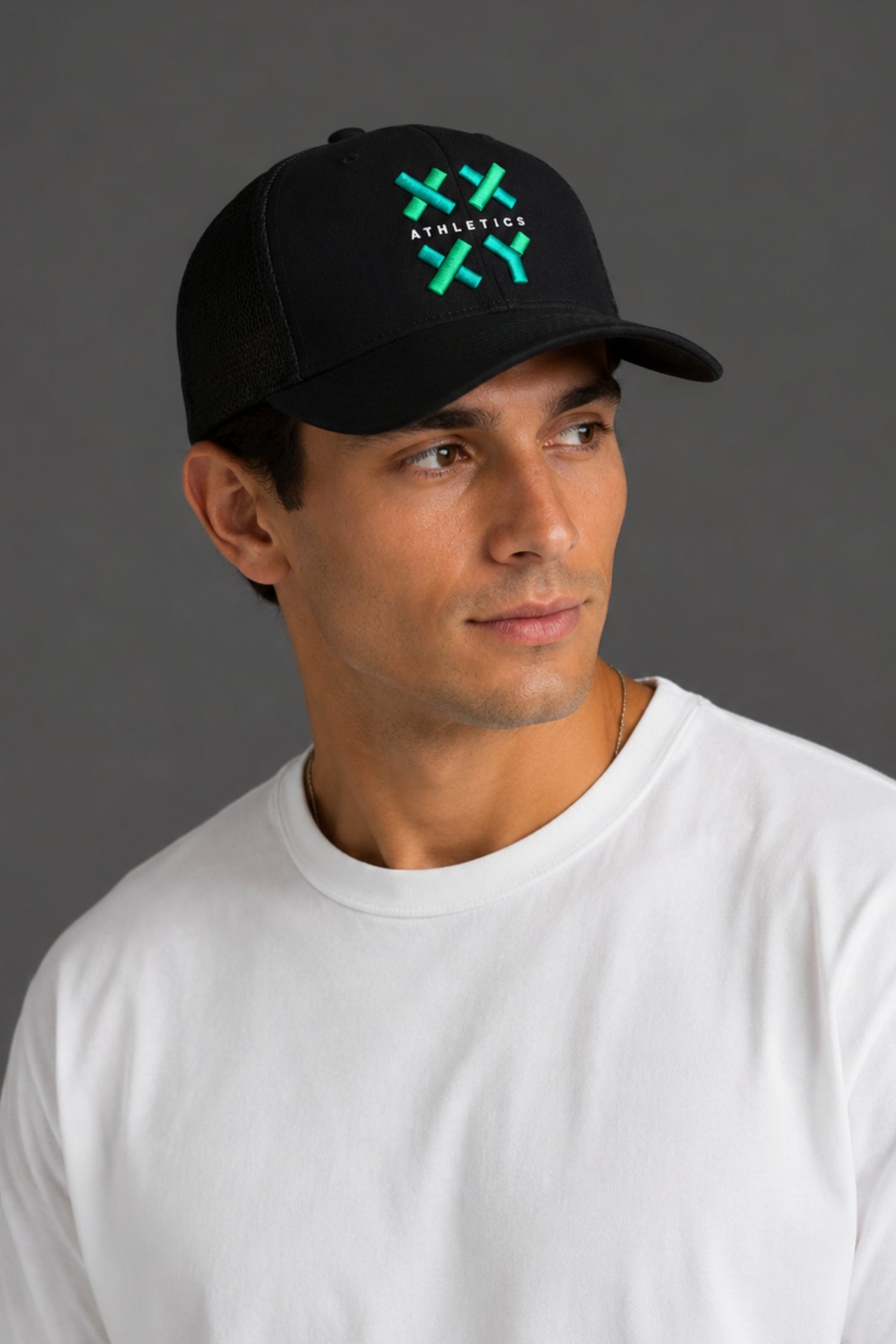 A man wearing a black baseball cap with a green and white embroidered logo that reads 'ATHLETICK' and 'XX', paired with a plain white t-shirt, set against a solid dark gray background.