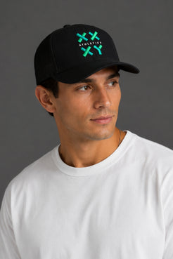 A man wearing a black baseball cap with a green and white embroidered logo that reads 'ATHLETICK' and 'XX', paired with a plain white t-shirt, set against a solid dark gray background. || Black with Color Logo