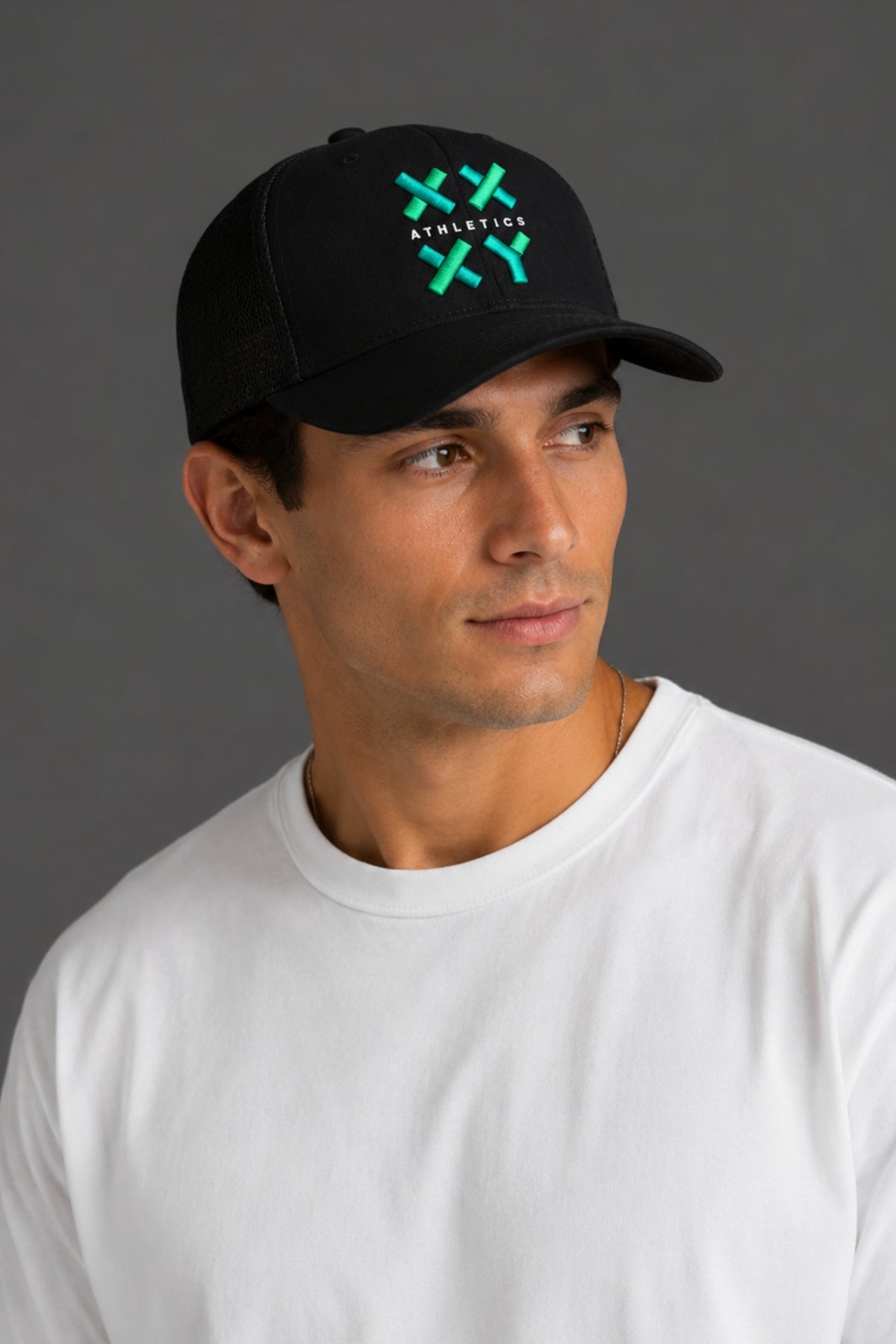 A man wearing a black baseball cap with a green and white embroidered logo that reads 'ATHLETICK' and 'XX', paired with a plain white t-shirt, set against a solid dark gray background. || Black with Color Logo