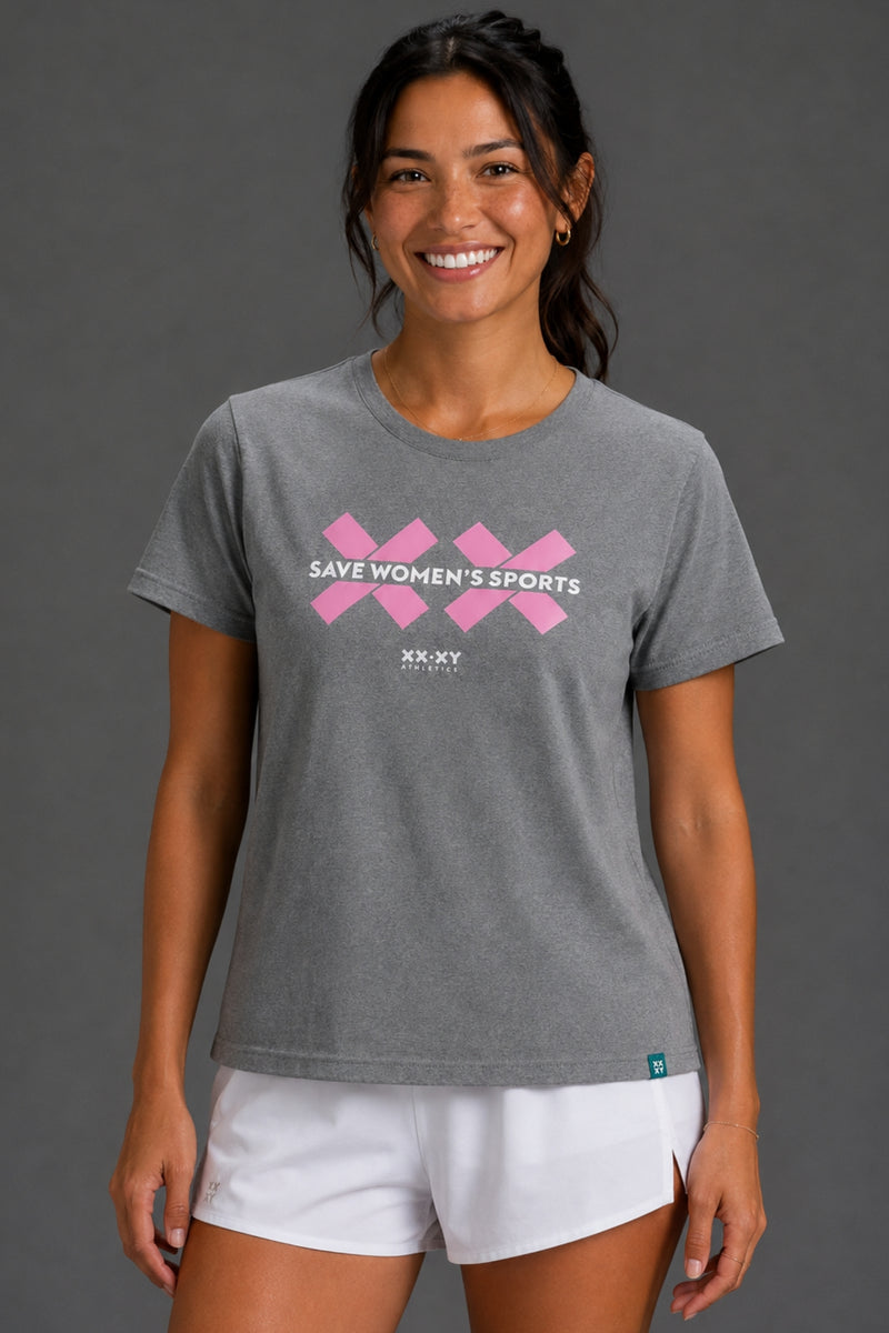 A woman wearing a heather gray short-sleeve t-shirt with a pink graphic that reads 'SAVE WOMEN'S SPORTS' and 'XX-XY SPORTS' underneath, paired with white athletic shorts, against a solid gray studio background. || Heather Gray