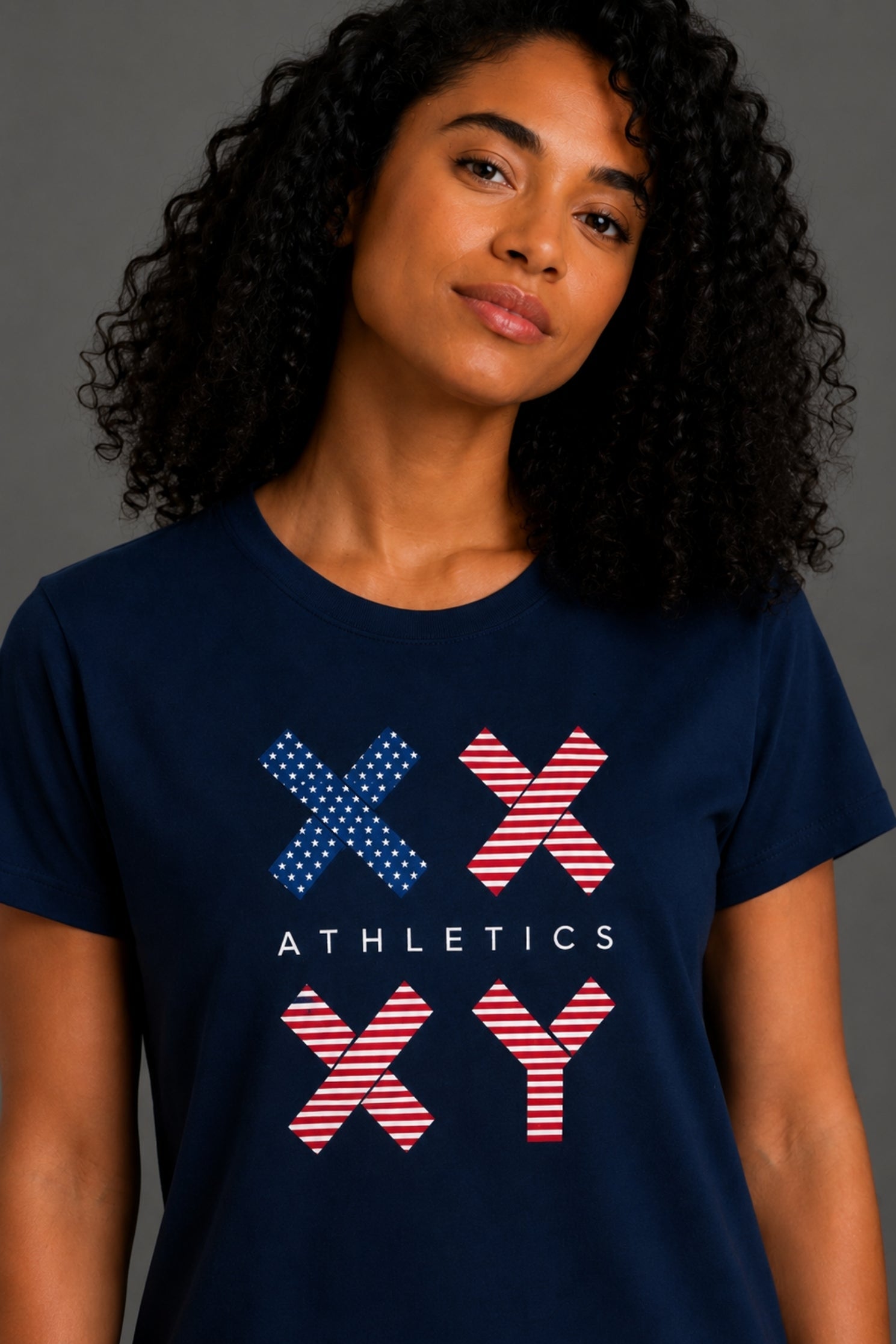 A woman with curly black hair wearing a navy blue short-sleeve t-shirt with a red, white, and blue graphic on the front against a gray studio background. || Dark Navy