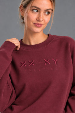 Close-up of burgundy XX-XY Athletics crewneck featuring tonal embroidered chest logo. || Red Wine