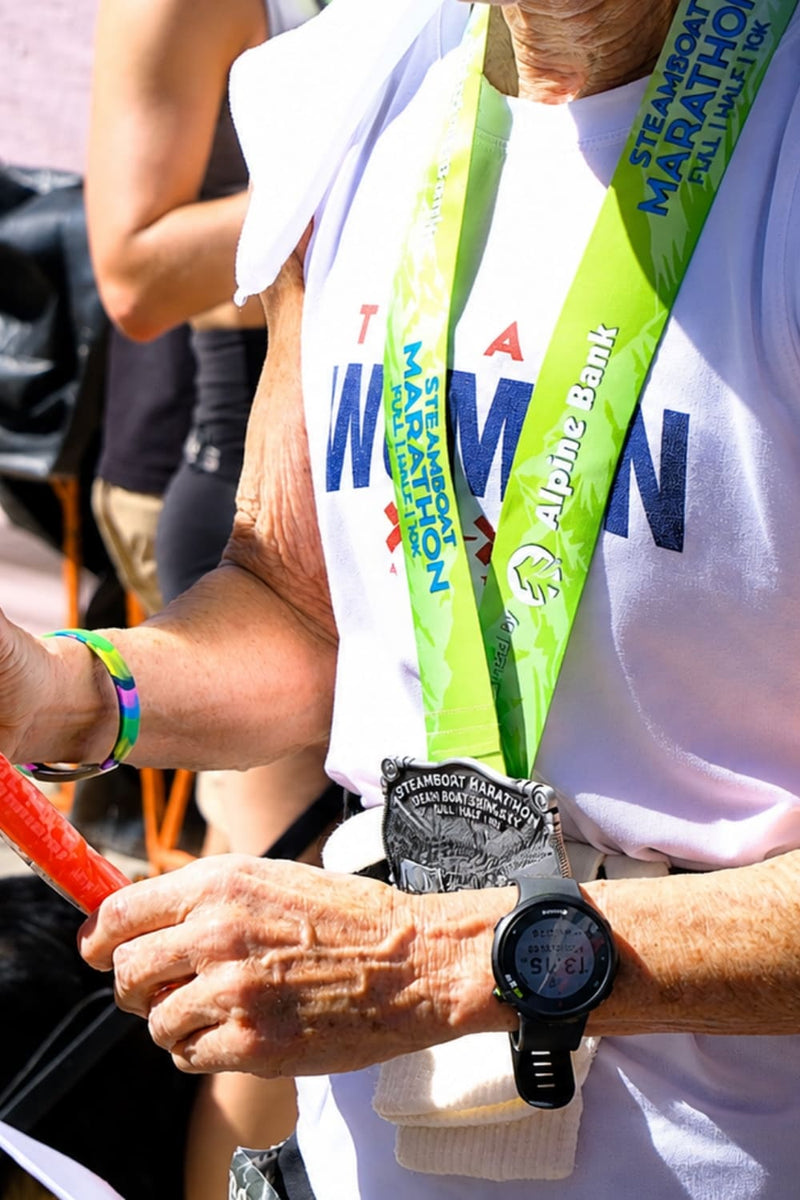 Close-up of a person at a marathon wearing a white t-shirt, a green and white race bib, a black digital watch, and a black medal, with another person partially visible in the background.