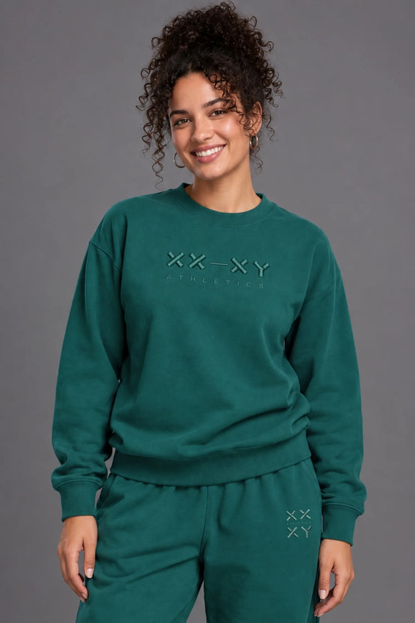 Woman wearing a teal XX-XY Athletics crewneck sweatshirt and matching sweatpants against a gray studio background. || Pine