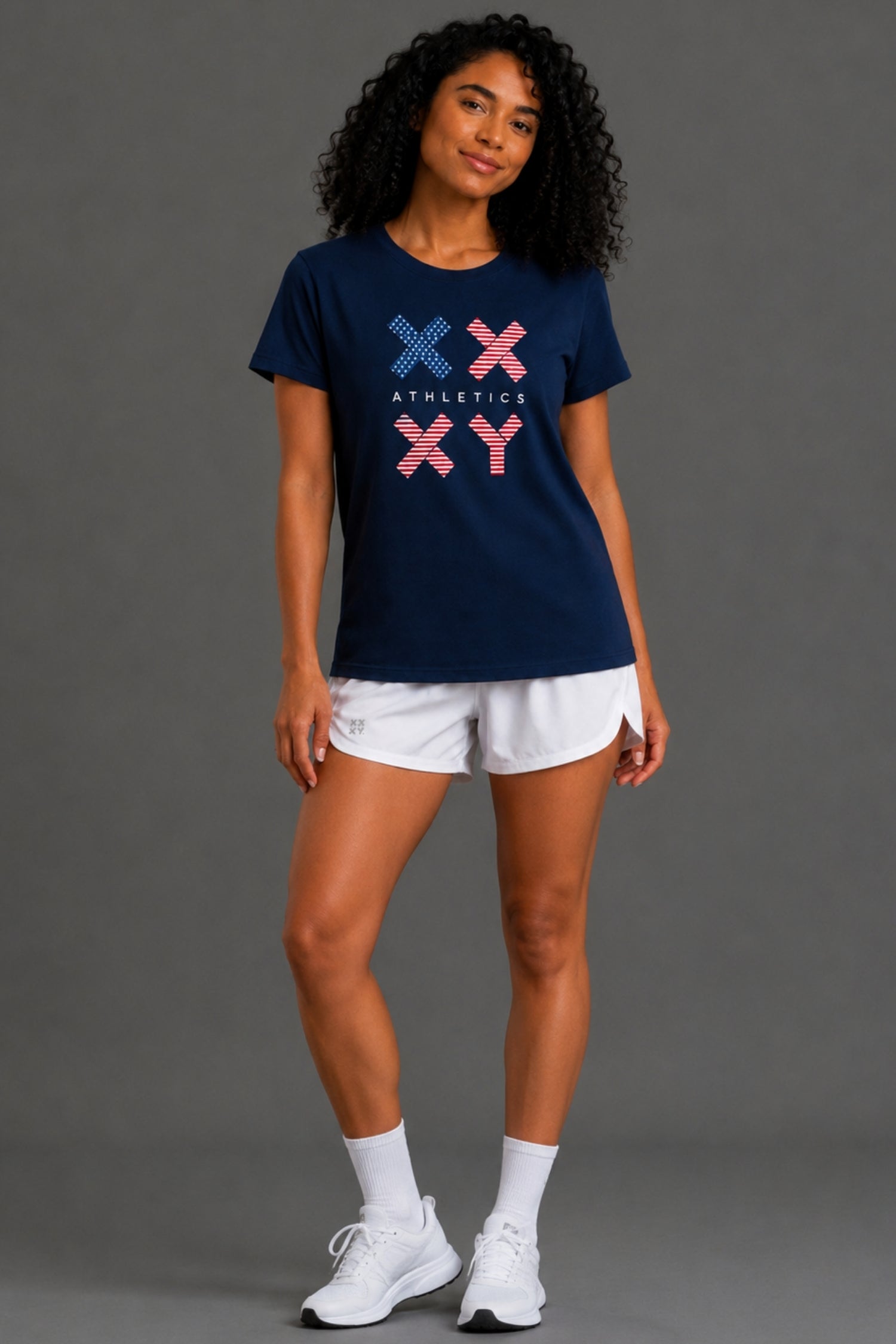 A woman with curly black hair wearing a navy blue short-sleeve t-shirt with a red, white, and blue graphic on the front, paired with white shorts, against a gray studio background. || Dark Navy