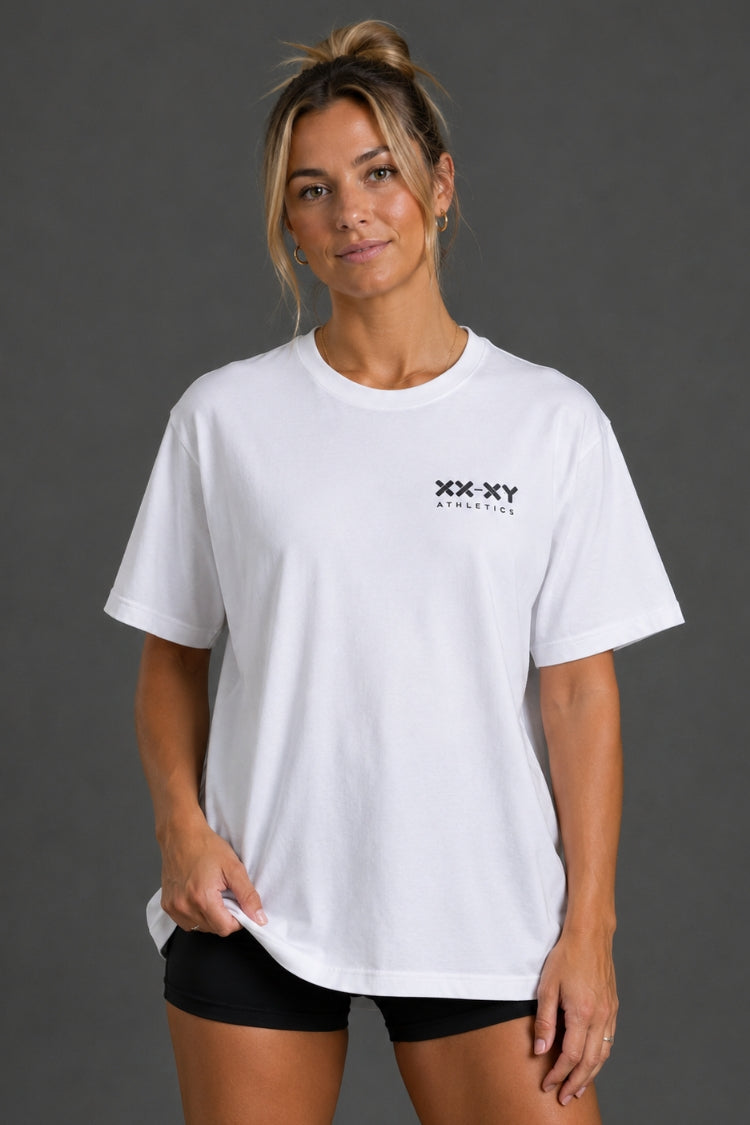 A woman modeling a white short-sleeve crewneck t-shirt with a small black 'XX XY ATHLETICS' logo on the left chest, paired with black athletic shorts against a dark gray studio background.