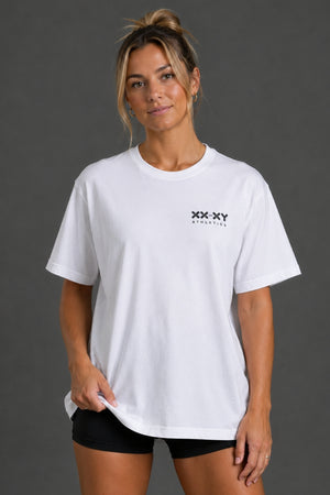 A woman modeling a white short-sleeve crewneck t-shirt with a small black 'XX XY ATHLETICS' logo on the left chest, paired with black athletic shorts against a dark gray studio background. || White
