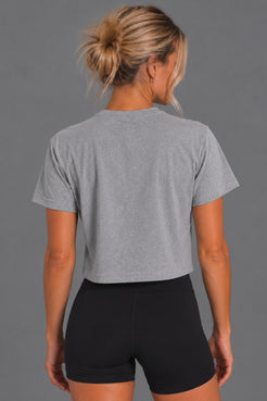Back view of blonde woman wearing the gray cropped Stand Up Save Women’s Sports tee with black bike shorts || Heather Gray