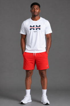 A man modeling a white short-sleeve t-shirt with a navy and red graphic logo on the chest, paired with bright red athletic shorts, white socks, and white sneakers, set against a solid gray studio background. || White