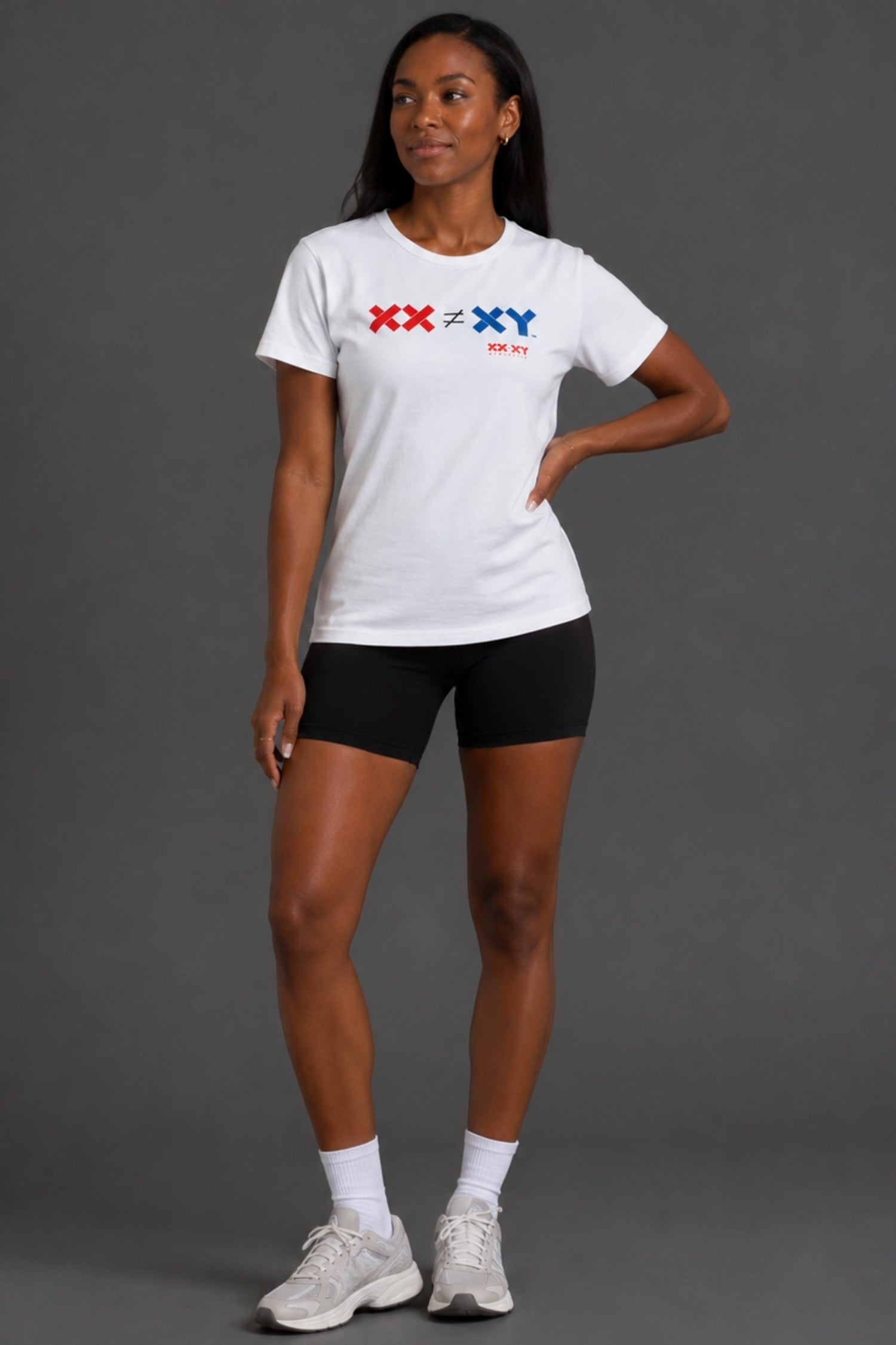 A woman modeling a white short-sleeve athletic T-shirt with a red and blue 'XX < XY > XX' graphic on the chest, paired with black bike shorts, white socks, and white sneakers against a dark gray studio background. || White
