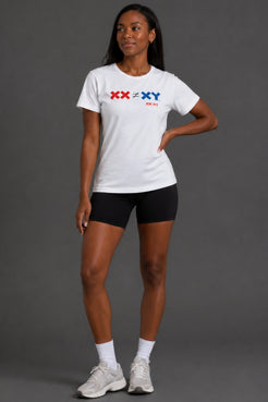A woman modeling a white short-sleeve athletic T-shirt with a red and blue 'XX < XY > XX' graphic on the chest, paired with black bike shorts, white socks, and white sneakers against a dark gray studio background. || White