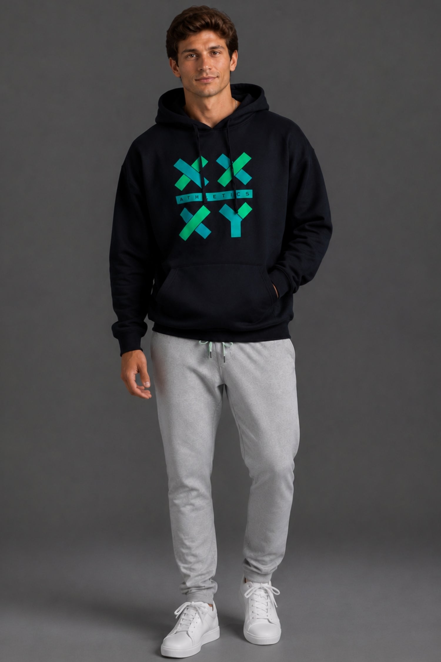 A man modeling a black pullover hoodie with a green and blue geometric 'X' and 'Y' graphic on the chest, paired with light gray sweatpants and white sneakers, against a dark gray studio background.