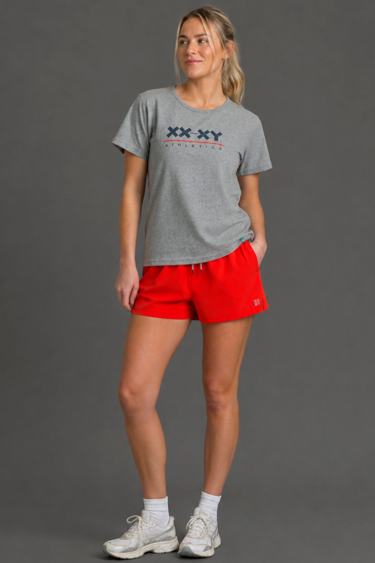 A woman modeling a heather gray t-shirt with a navy and red 'XX XY ATHLETICS' graphic on the chest, paired with bright red athletic shorts, white socks, and white sneakers, set against a solid gray studio background.