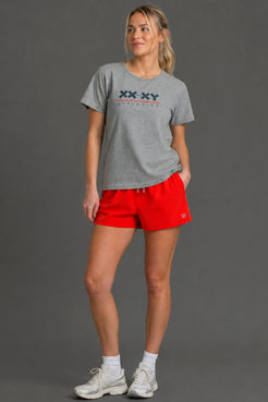 A woman modeling a heather gray t-shirt with a navy and red 'XX XY ATHLETICS' graphic on the chest, paired with bright red athletic shorts, white socks, and white sneakers, set against a solid gray studio background. || Light Heather Gray