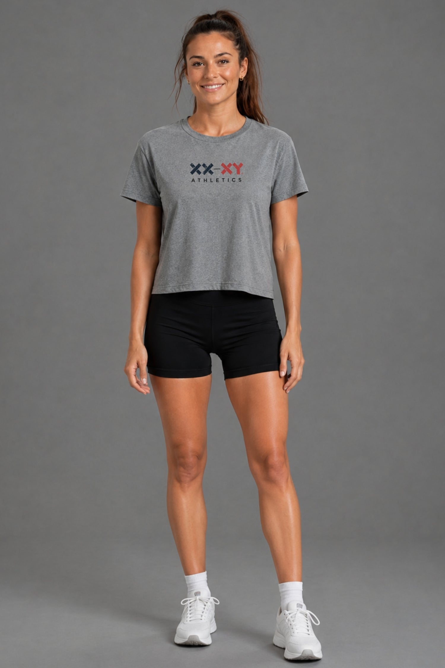 A woman modeling a heather gray cropped t-shirt with a 'XX-XY ATHLETICS' logo on the chest, paired with black athletic shorts, white socks, and white sneakers, against a solid gray studio background. || Heather Gray