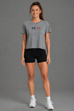 A woman modeling a heather gray cropped t-shirt with a 'XX-XY ATHLETICS' logo on the chest, paired with black athletic shorts, white socks, and white sneakers, against a solid gray studio background. || Heather Gray