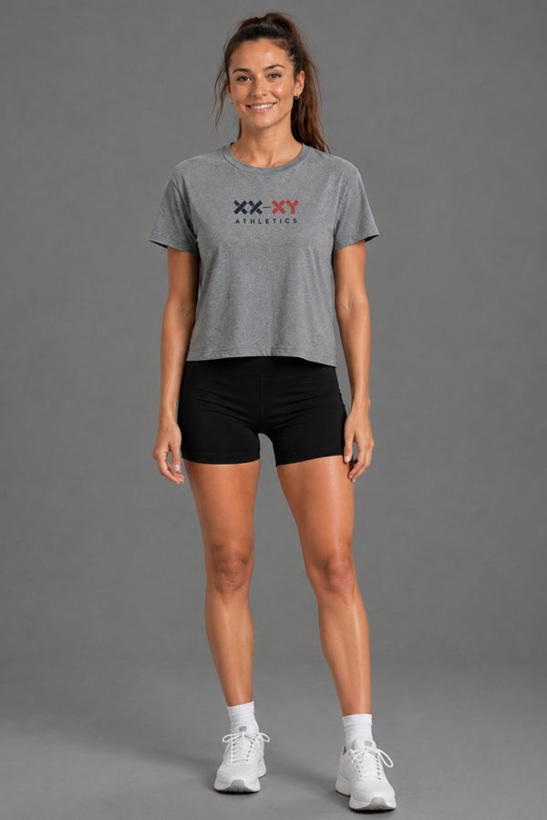 A woman modeling a heather gray cropped t-shirt with a 'XX-XY ATHLETICS' logo on the chest, paired with black athletic shorts, white socks, and white sneakers, against a solid gray studio background. || Heather Gray