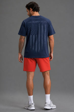 A man viewed from the back wearing a navy blue t-shirt with a distressed American flag graphic and the words 'SAVE WOMEN'S SPORTS' across the upper back, paired with red athletic shorts, white socks, and gray sneakers, against a solid gray studio background. || Navy