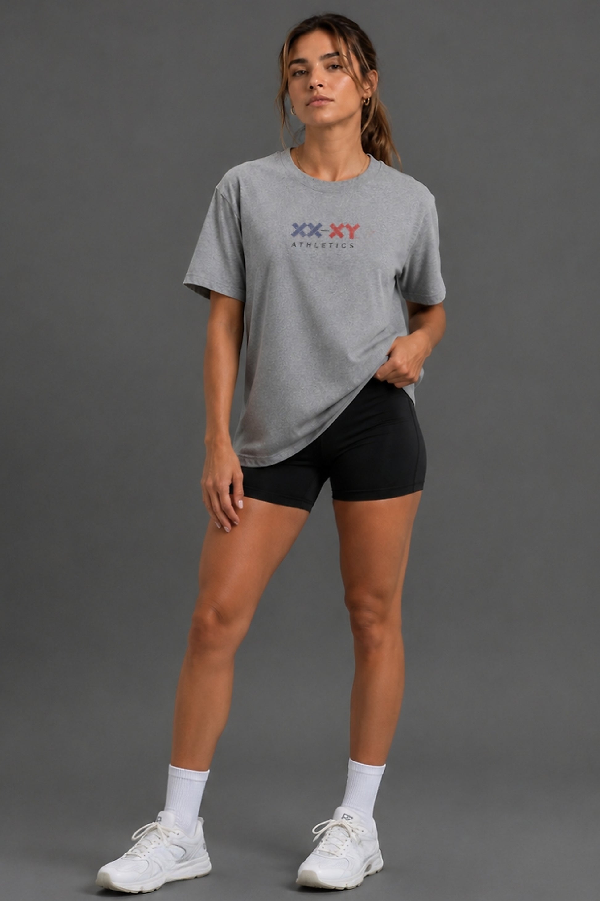 A woman modeling a heather gray short-sleeve athletic t-shirt with a red and blue 'XX-XY ATHLETICS' logo on the chest, paired with black athletic shorts, white socks, and white sneakers against a solid gray studio background.