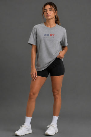 A woman modeling a heather gray short-sleeve athletic t-shirt with a red and blue 'XX-XY ATHLETICS' logo on the chest, paired with black athletic shorts, white socks, and white sneakers against a solid gray studio background. || Heather Gray